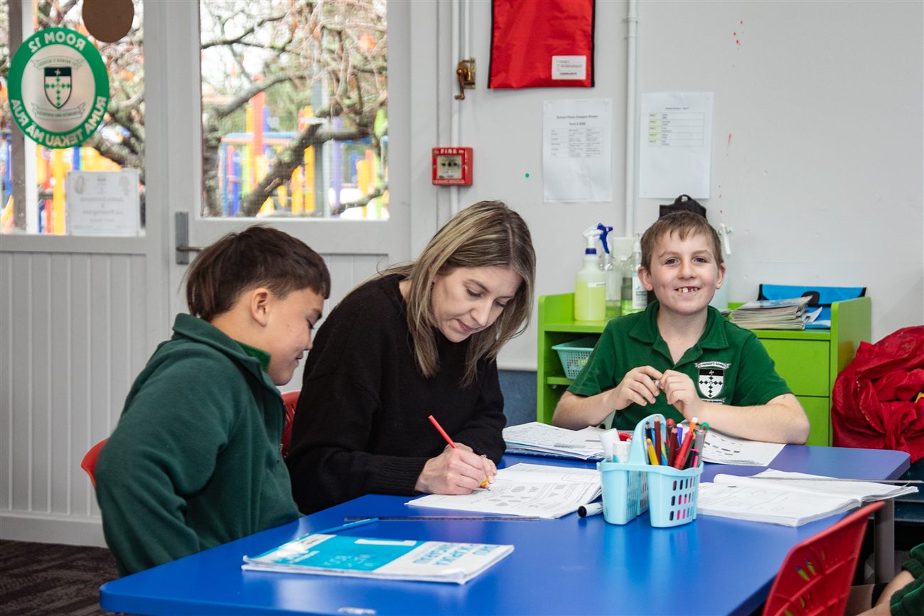 A teacher working alongside two students at a classroom table, one of them smiling at the camera