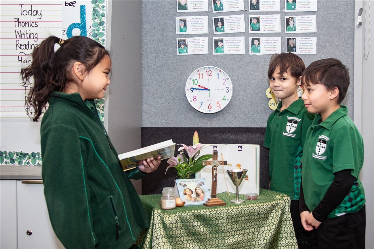 Two boys and a girl standing around a classroom prayer table with a cross, a candle and a chalice