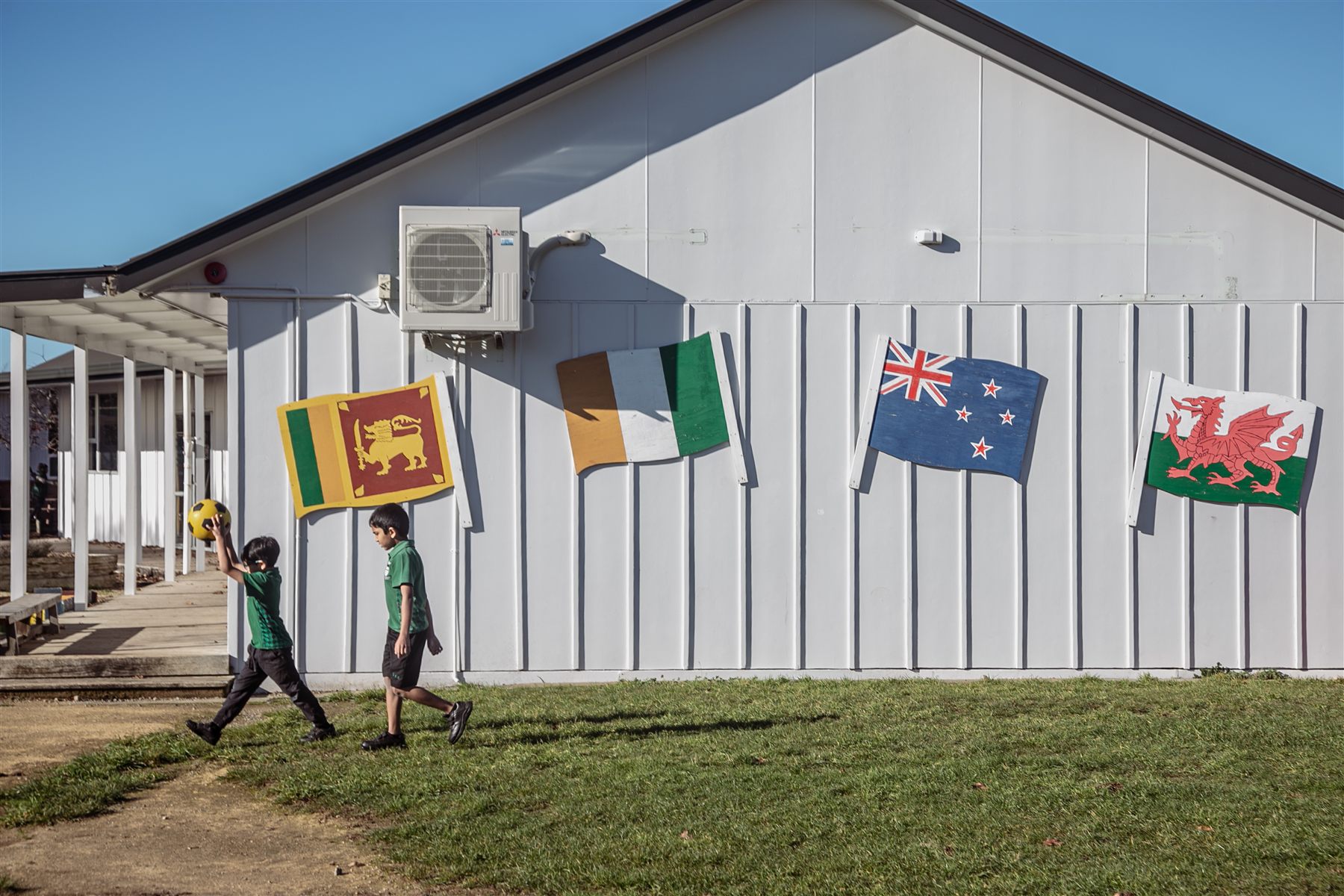 Two boys playing with a yellow ball in front of a school building decorated with flags from around the world