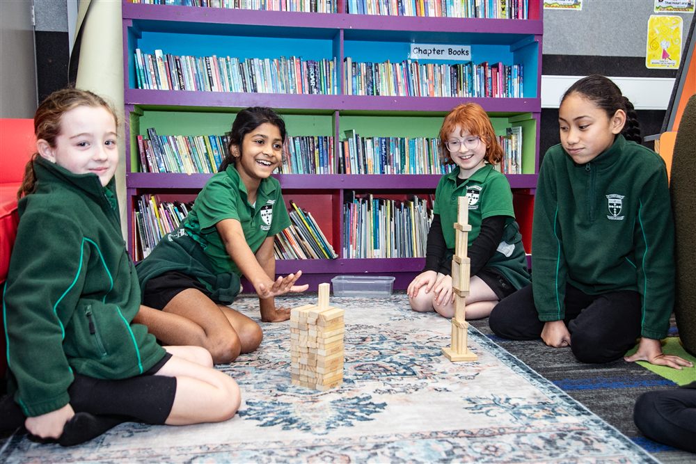 Four students concentrating as they build a tall tower of wooden blocks on the library carpet